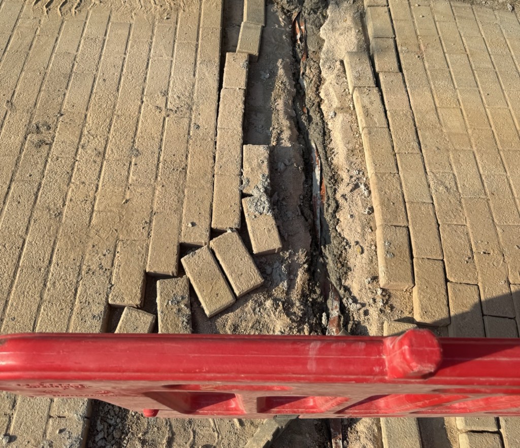 Εικόνα ενός πεζοδρομίου με σπασμένα κυβόλιθους και μία κόκκινη μπάρα ασφαλείας μπροστά.